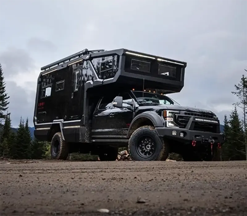Black overland expedition truck with a custom camper parked on a mountain road under a cloudy sky.