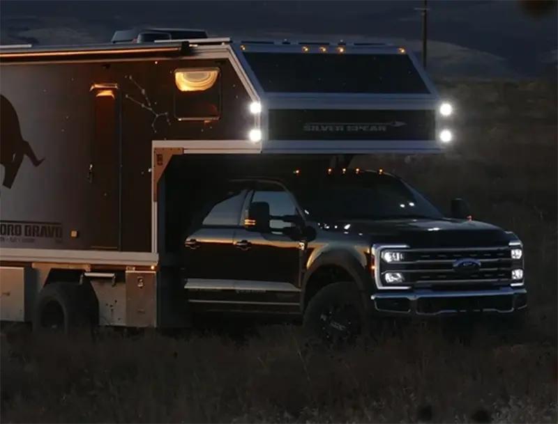 Off-road truck with camper trailer parked at dusk with headlights on in a rugged outdoor setting.