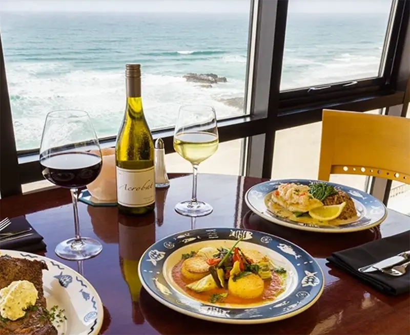 Oceanfront dining table with wine, seafood entrees, and views of the waves through large restaurant windows.